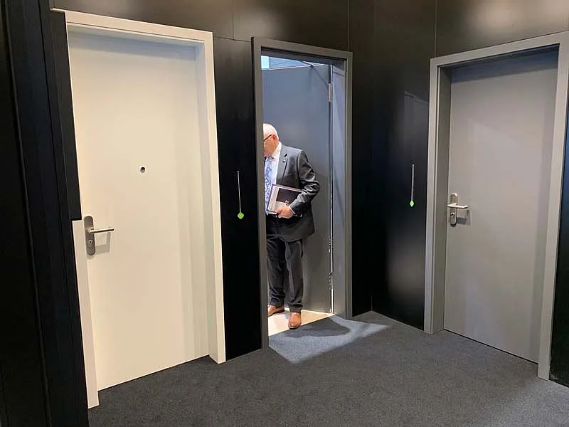 Drei Türöffnungen in schwarzer Messewand: links geschlossene weiße Tür mit Türspion und Griff; mittlere graue Tür geöffnet, ein Mann im Anzug mit Unterlagen tritt hindurch; rechts geschlossene graue Tür mit Griff und Schloss; dunkler Teppichboden.